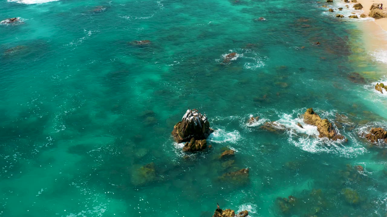 Aerial frame over clear turquoise water at Dreams Beach, with the main rock lower center, surf curling around smaller rocks, sand at upper right, and no visible people.