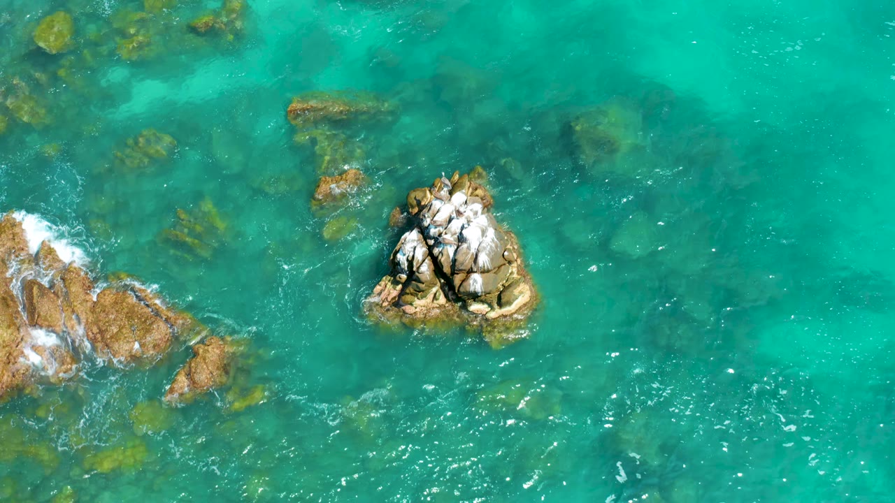 Aerial view over Dreams Beach water shows the main cracked rock slightly right of center, submerged rocks below the surface, small white wave flecks, and no visible people.