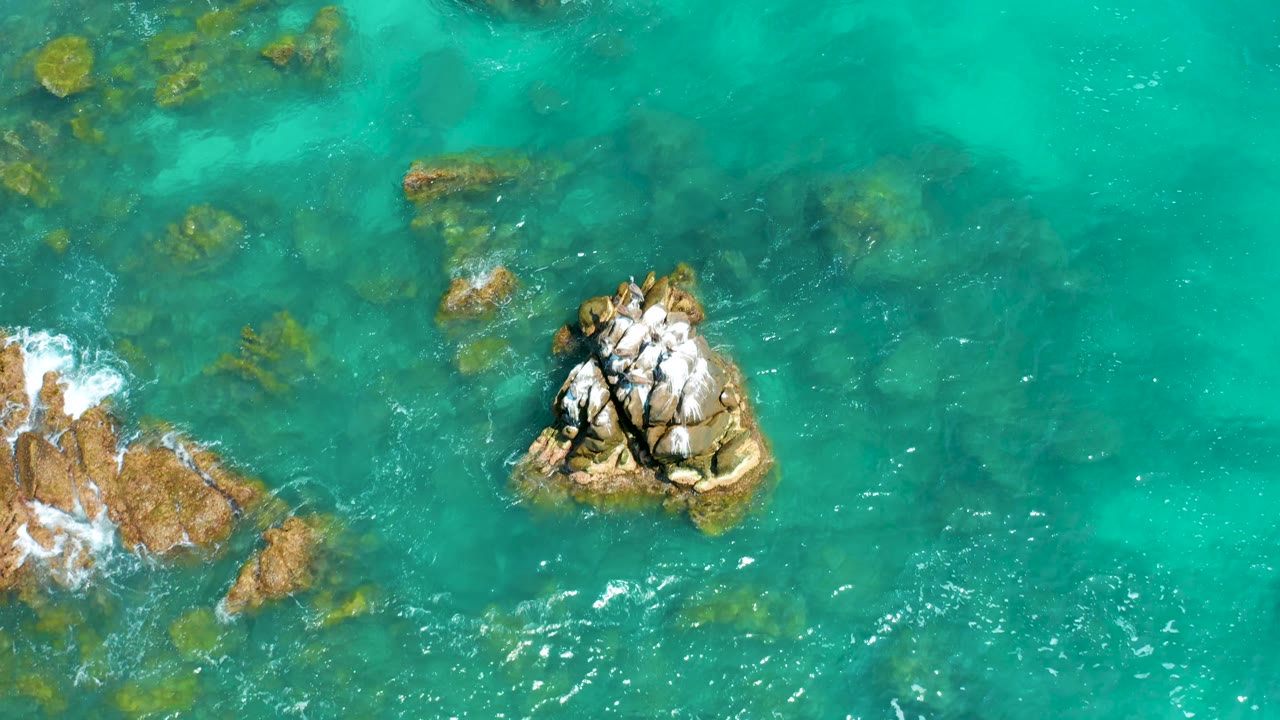 Overhead frame from Dreams Beach shows the cracked rock outcrop farther right, bright turquoise water across the frame, foam brushing rocks on the left edge, and no visible people.