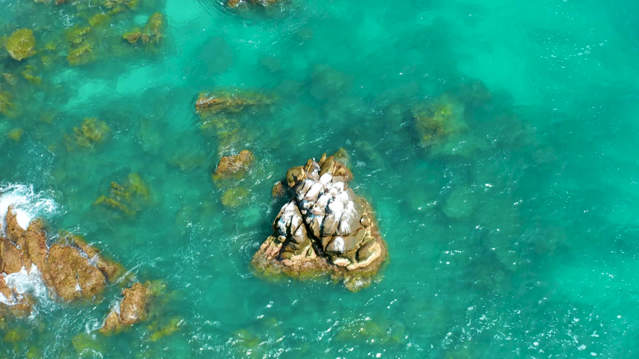 At Dreams Beach, the aerial composition places the main cracked rock below center, with blue-green water, faint submerged rocks, left-edge foam, and no visible people.