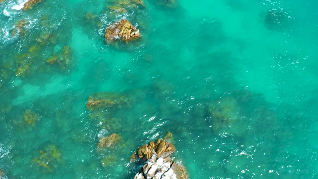 Overhead view of turquoise water at Dreams Beach in Baja California Sur, Mexico, with several submerged rocks, two exposed rock clusters, white foam at the edges, and no visible people.