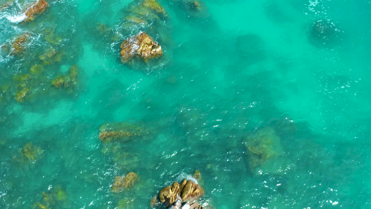 Top-down view of turquoise sea at Dreams Beach in Baja California Sur, Mexico, with exposed rocks near the top and lower frame, faint white foam, darker underwater patches, and no visible people.
