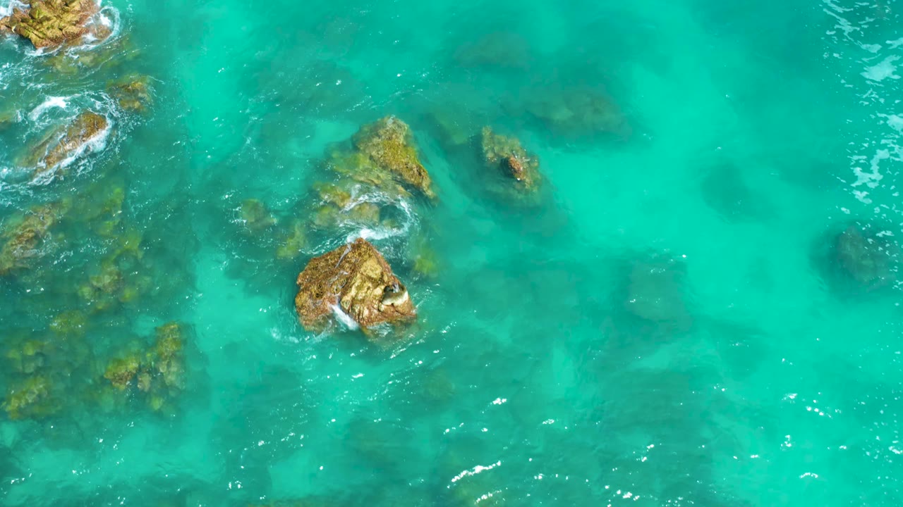 Aerial view at Dreams Beach in Baja California Sur, Mexico, with the main tan rock near center, smaller rocks above it, and clear turquoise water extending to the right with no visible people.