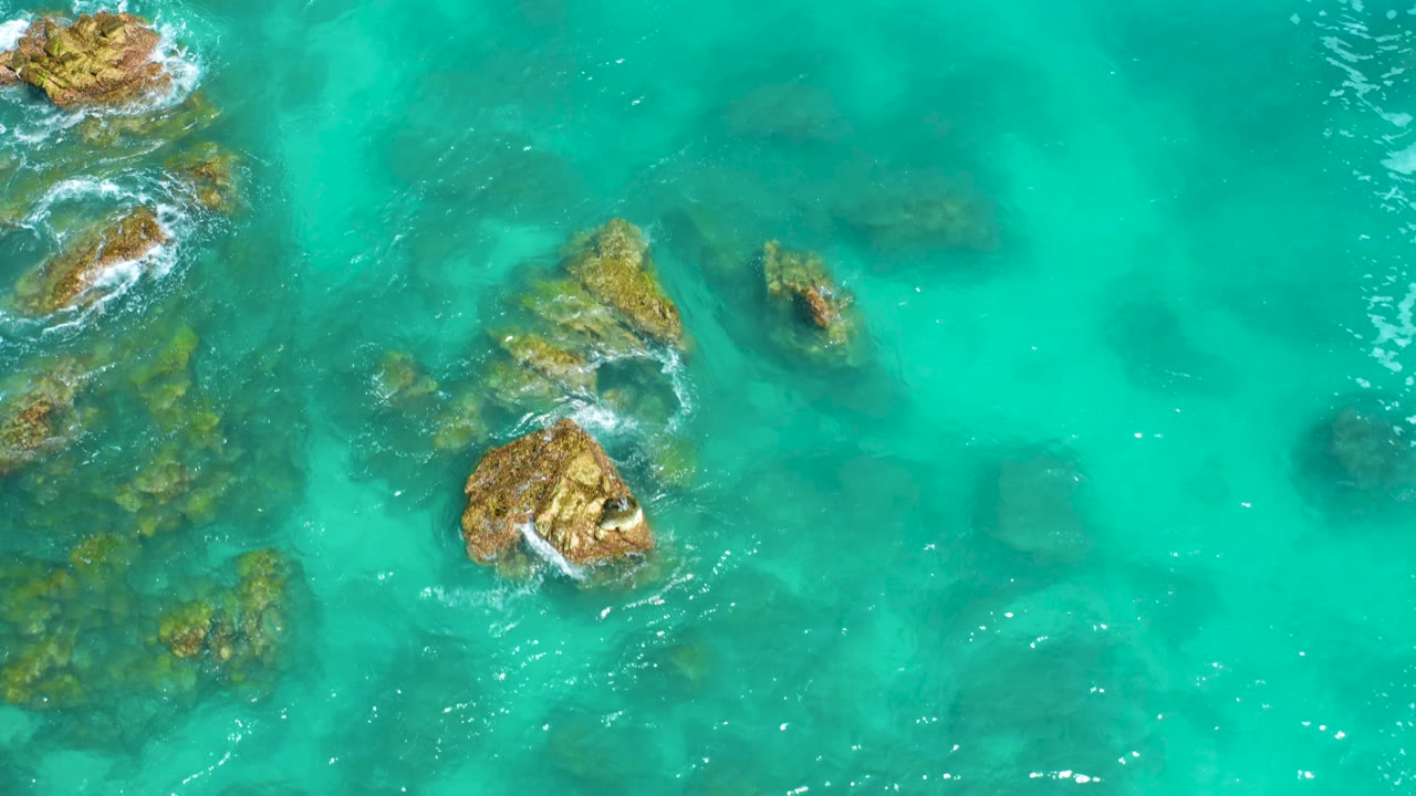 Top-down aerial frame from Dreams Beach in Baja California Sur, Mexico, showing tan rocks grouped from the upper left toward center with blue-green water and faint submerged shapes, no visible people.
