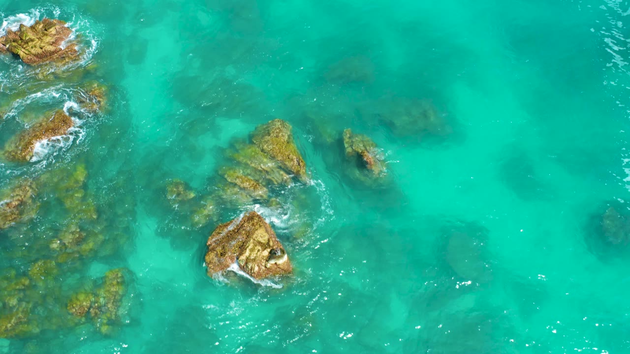 Aerial frame at Dreams Beach in Baja California Sur, Mexico, with rocks along the left and lower center, small white wave breaks, and open turquoise water across the right, no visible people.