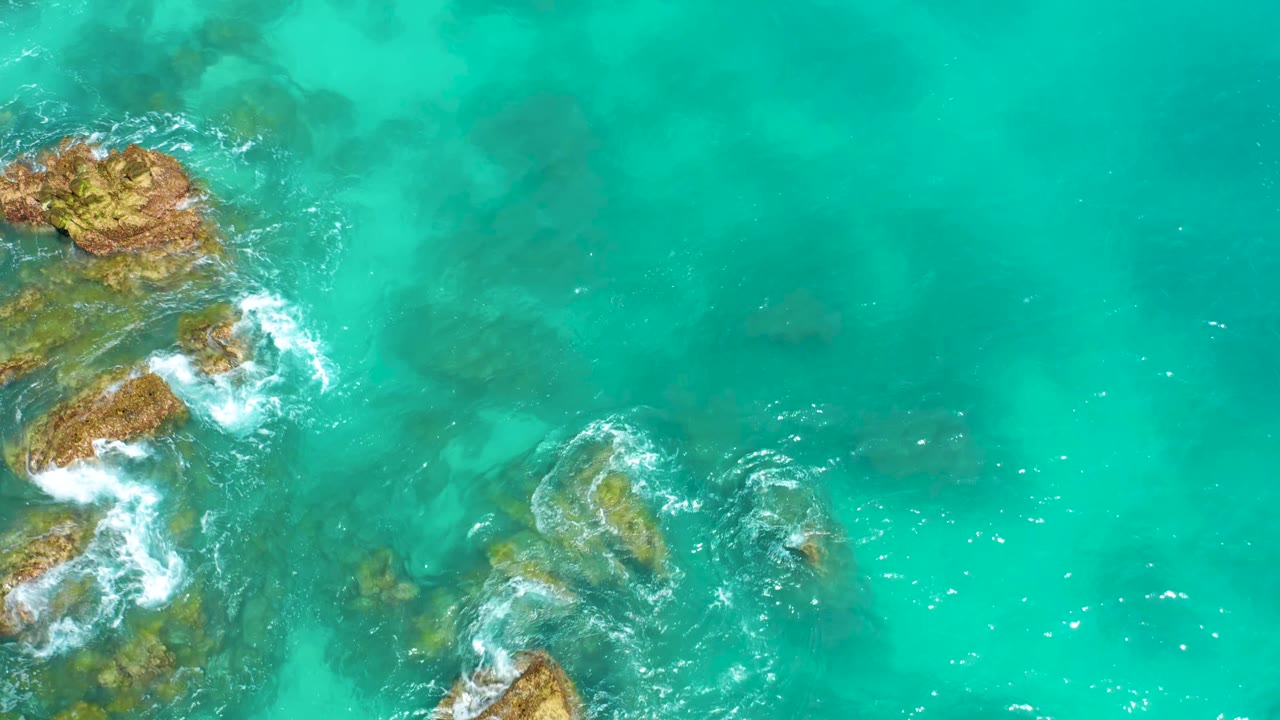 Aerial top view at Dreams Beach in Baja California Sur with exposed rocks near the left edge, pale surf foam, clear green-blue water, and no visible people.