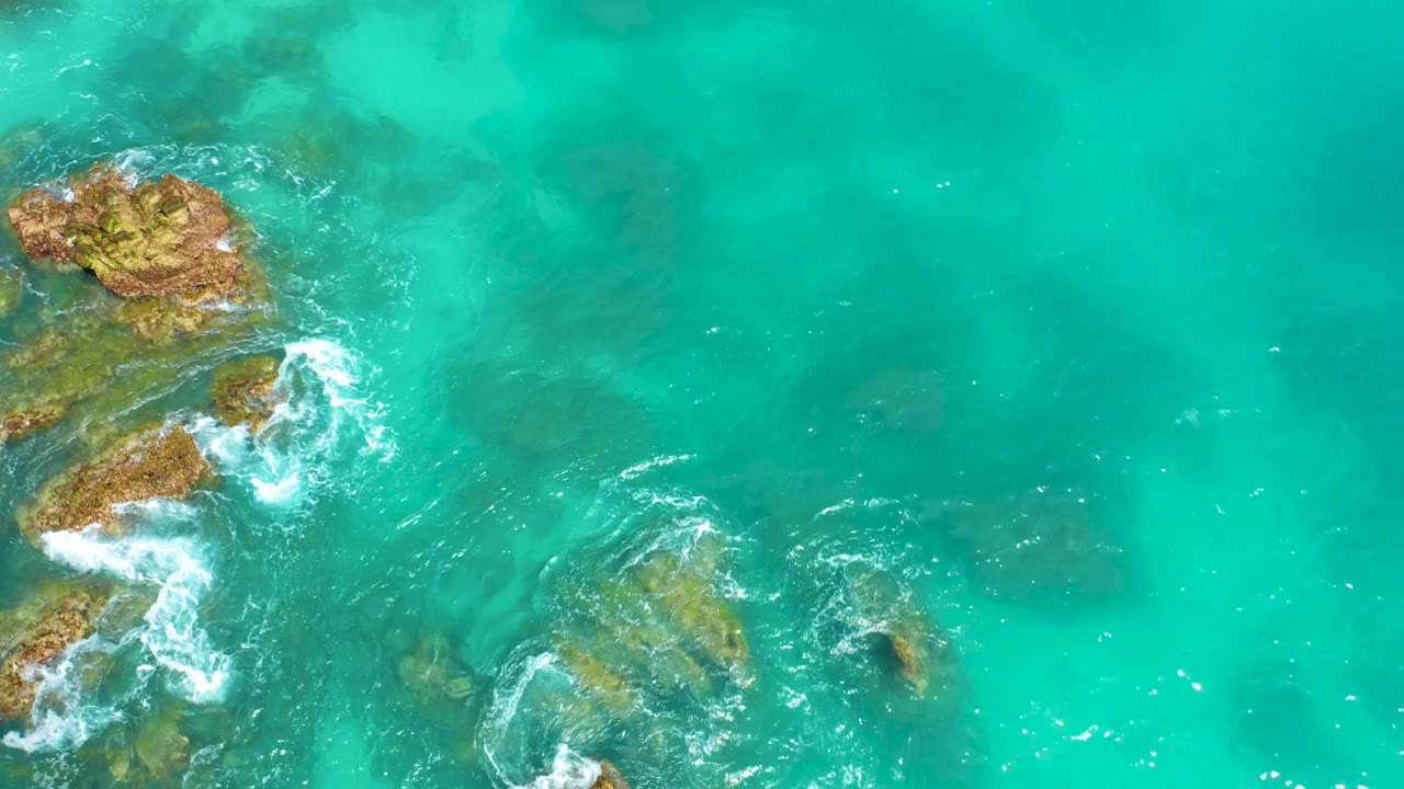 Top-down drone view at Dreams Beach in Baja California Sur showing clustered rocks in the upper left, smaller stones under clear water, breaking white waves, and no visible people.