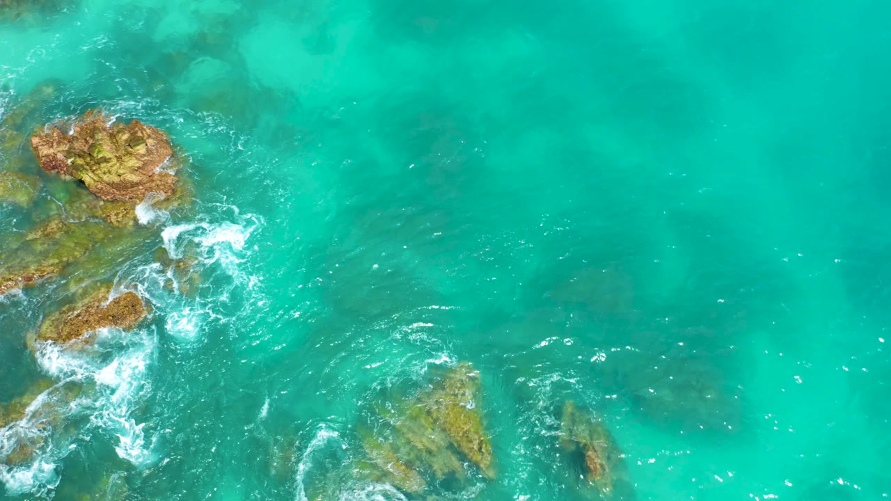 Overhead frame from Dreams Beach in Baja California Sur with rocks entering from the left, bright foam beside the stones, shallow turquoise water, and no visible people.