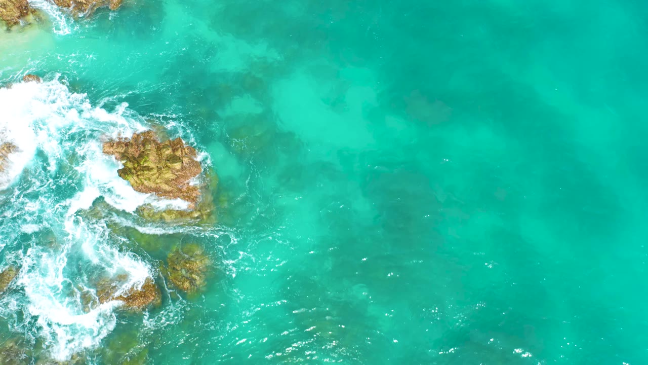 Aerial frame from Dreams Beach in Baja California Sur showing a central-left rock outcrop, white foam curling around it, open turquoise water to the right, and no visible people.