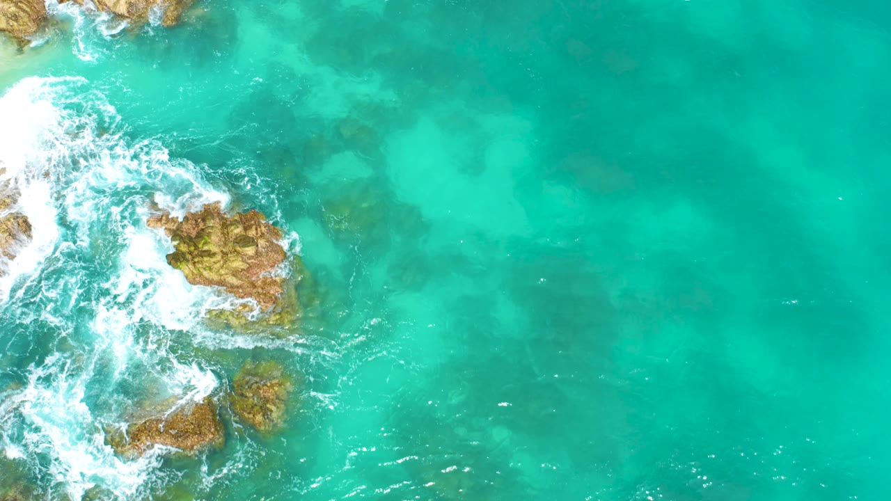 Top-down Dreams Beach frame with several tan rocks along the left edge, white surf spreading outward, clear turquoise water across the right half, and no visible people.
