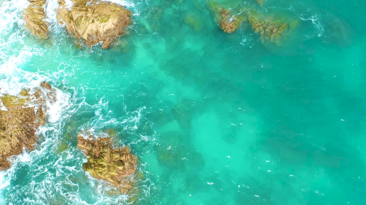 Overhead frame from Dreams Beach, Baja California Sur, with rocky outcrops along the left and top edges, clear blue-green water across the center, and no visible people.