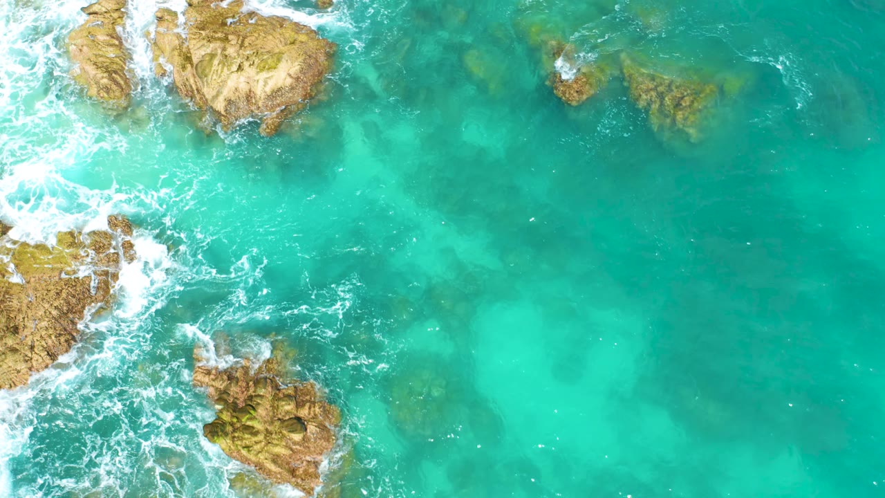 At Dreams Beach, Baja California Sur, white surf pushes against clustered tan rocks on the left while turquoise water opens across the right side, with no visible people.