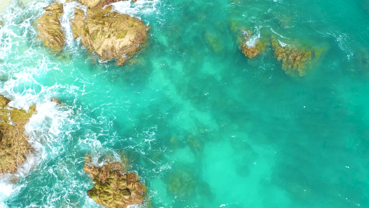 Overhead view at Dreams Beach, Baja California Sur, showing clear turquoise ocean, tan rocks partly surrounded by foam, and darker underwater patches, with no visible people.
