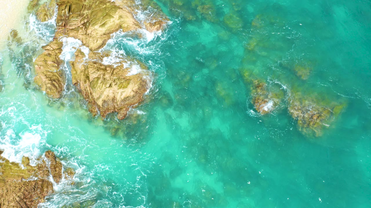 Overhead frame from Dreams Beach in Baja California Sur, Mexico, showing tan coastal rocks at left, turquoise water at right, white surf around the edges, and no visible people.