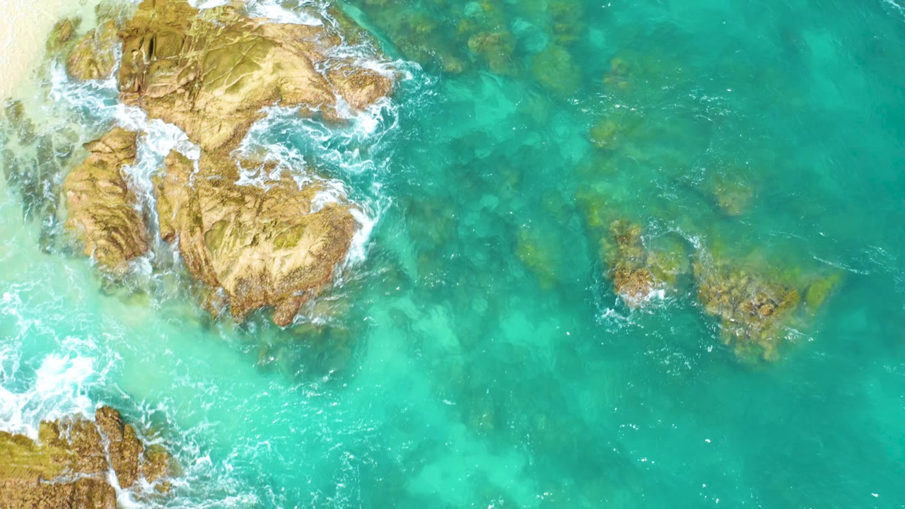 Overhead frame from Dreams Beach in Baja California Sur, Mexico, showing jagged tan rocks along the left, clear water across the middle, small reef patches to the right, and no visible people.