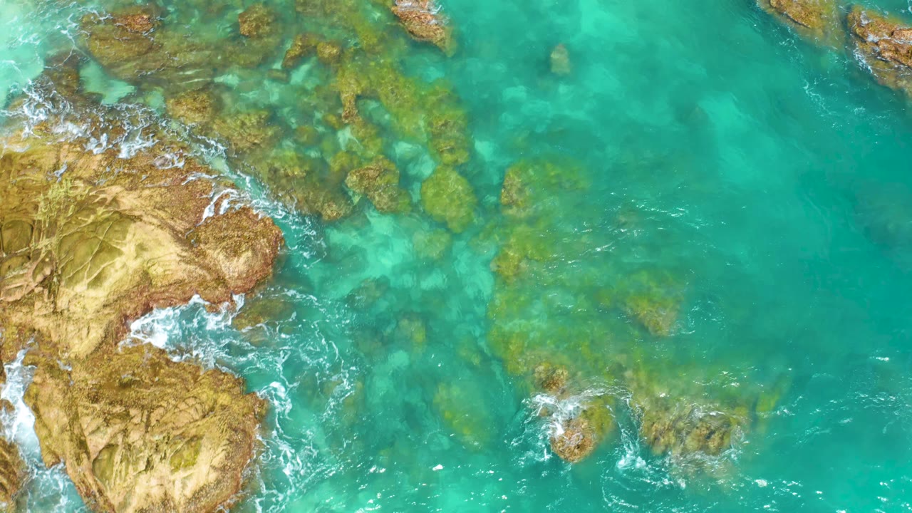 Aerial frame from Dreams Beach in Baja California Sur with clear turquoise water, submerged rock patches, tan shoreline rock at left, and no visible people.