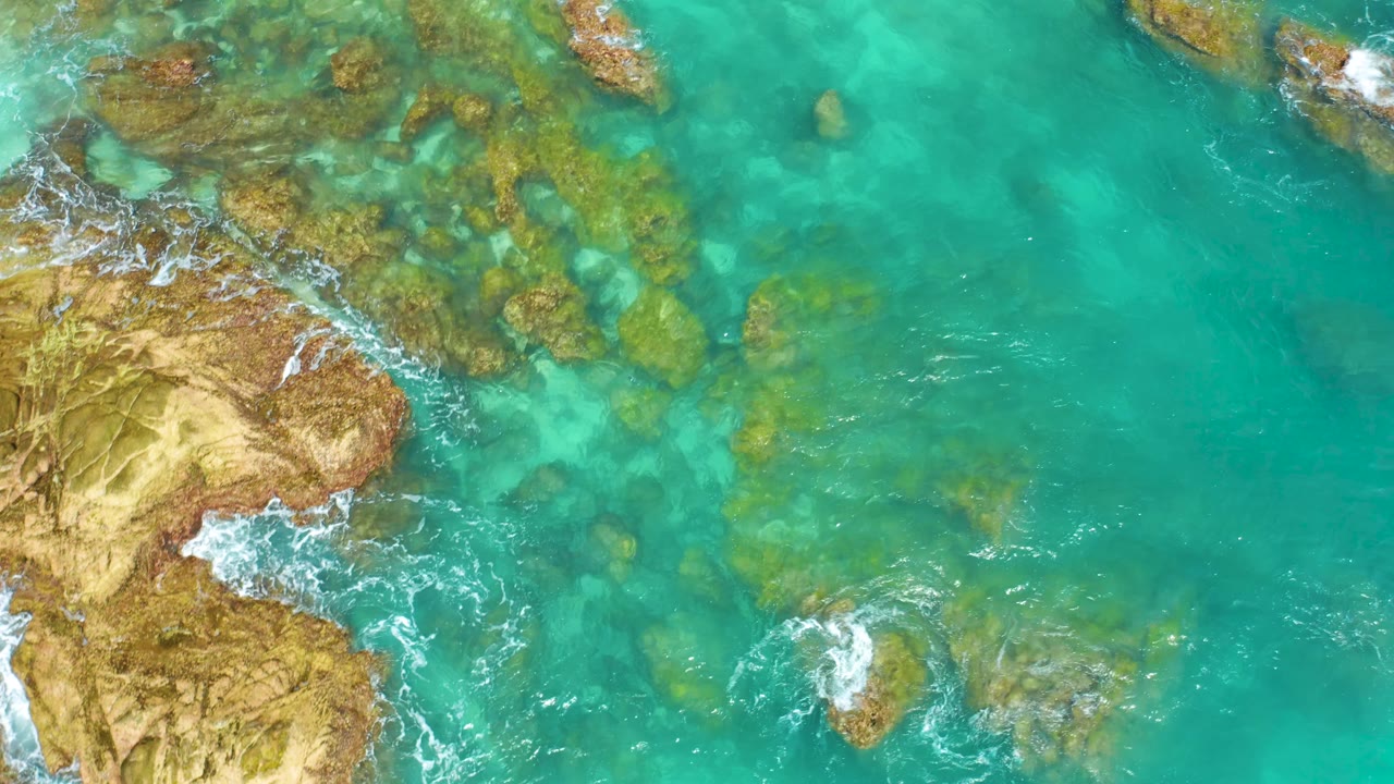Overhead coastal view at Dreams Beach in Baja California Sur with tan rocks along the left edge, bright turquoise water across the frame, scattered submerged rocks, and no visible people.