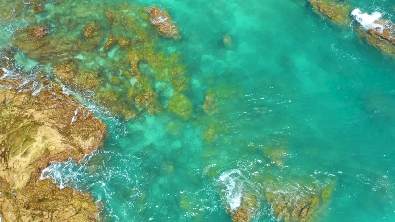 Straight-down view over Dreams Beach in Baja California Sur with turquoise water, rock outcrops near the left and lower areas, faint foam trails, and no visible people.