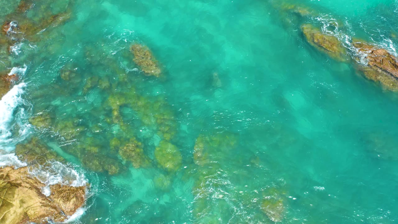 Aqua water at Dreams Beach in Baja California Sur shows a broad submerged rock patch near the lower left, smaller rocks near the top, sunlight on the surface, and no visible people.