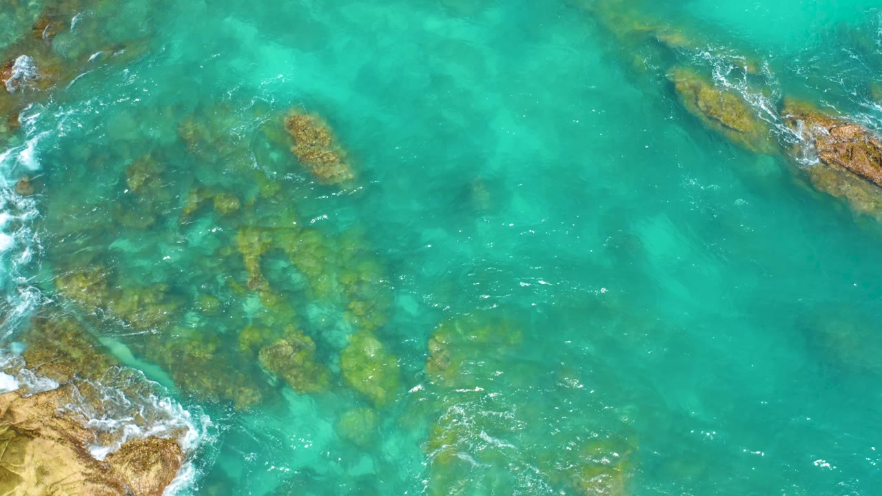 Top-down frame from Dreams Beach in Baja California Sur with green-brown rocks visible under turquoise water, a tan rocky edge at left, thin white foam, and no visible people.