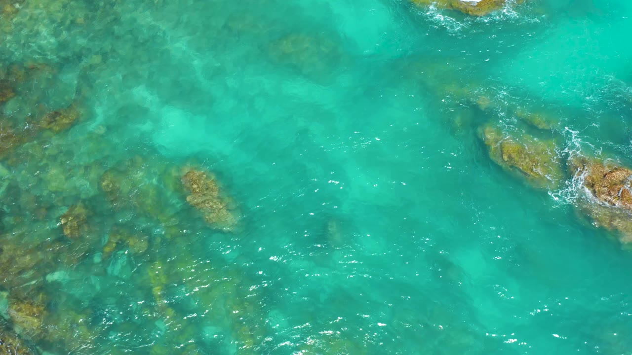 Overhead frame from Dreams Beach, Baja California Sur, with darker submerged rocks on the left, open turquoise water through the center, foam on a right-side rock, and no visible people.