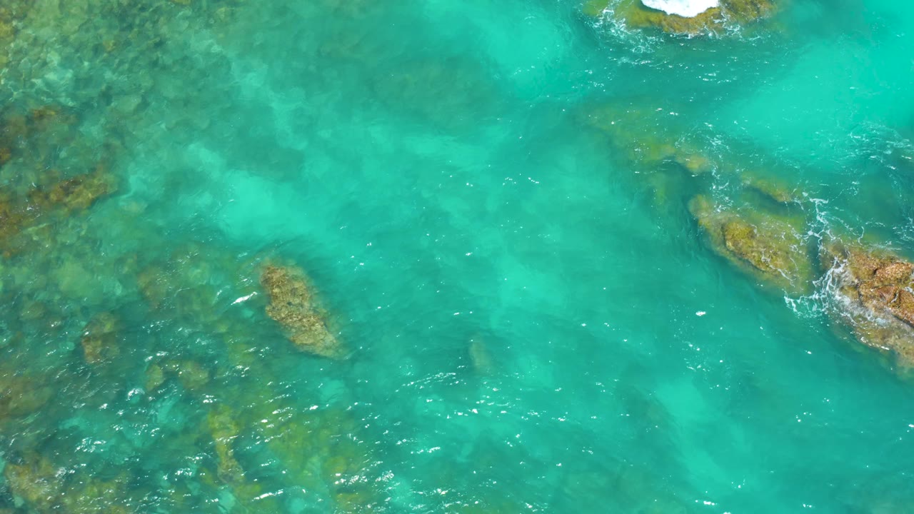 Aerial view at Dreams Beach in Baja California Sur with a broad turquoise channel, clustered submerged rocks at left, a small exposed rock near the top edge, and no visible people.
