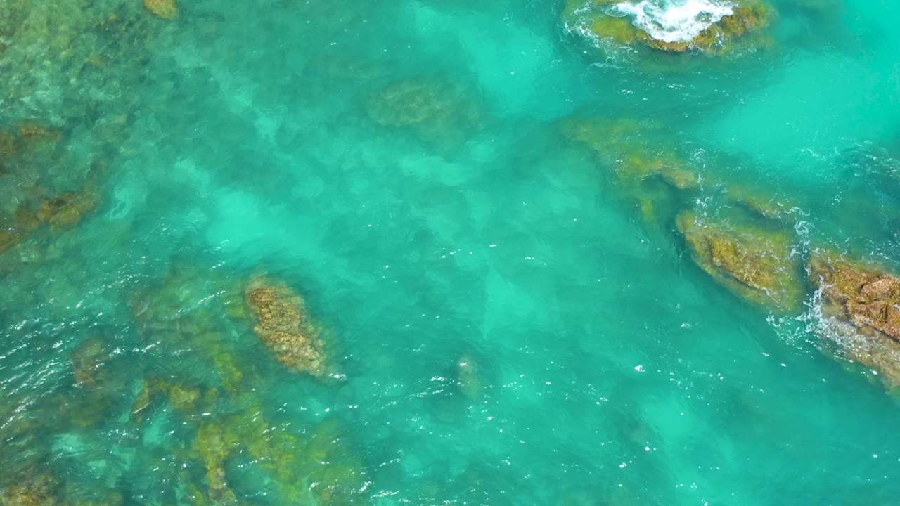 Top-down Dreams Beach frame in Baja California Sur showing scattered brown-green rocks under turquoise water, white surf touching rocks near the top and right edges, and no visible people.