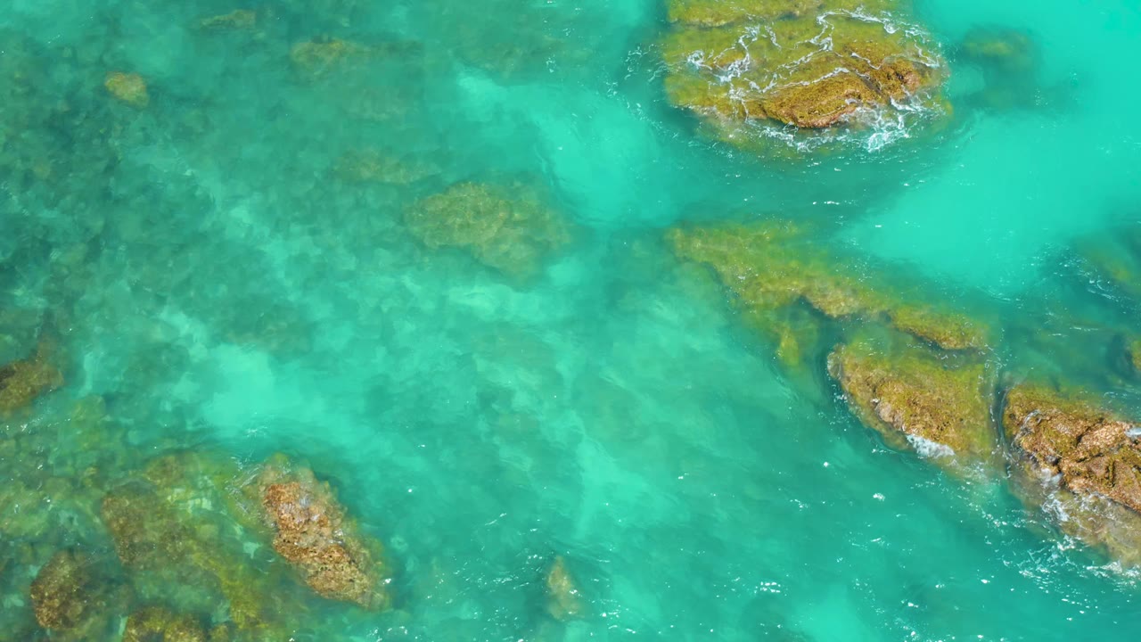 Drone-like overhead frame at Dreams Beach showing clear blue-green water, several submerged rocks, one larger exposed rock near the top, and no visible people.
