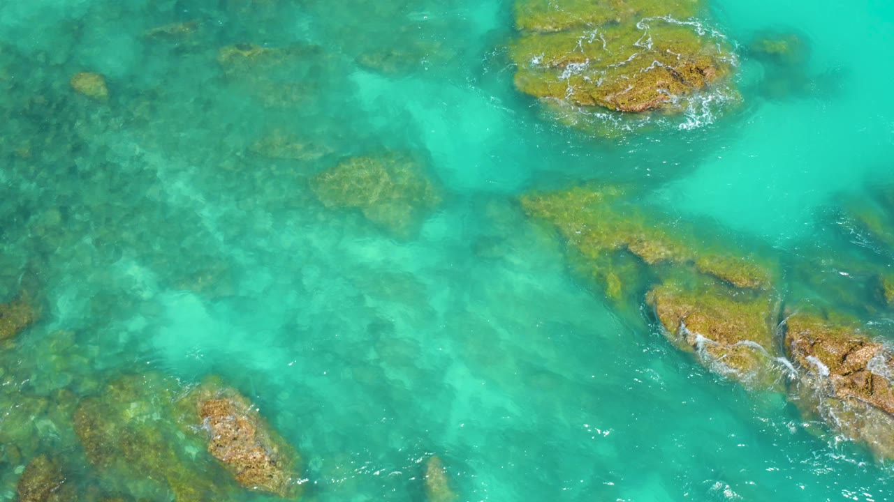 Aerial frame from Dreams Beach with turquoise shallows, darker underwater rock patches, exposed brown rocks at the upper and right edges, and no visible people.