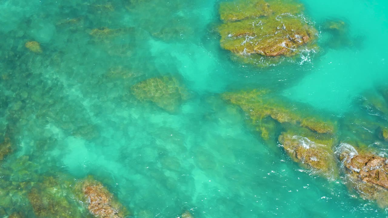 Top-down view at Dreams Beach with a large brown rock near the upper center, smaller submerged formations through green-blue water, and no visible people.