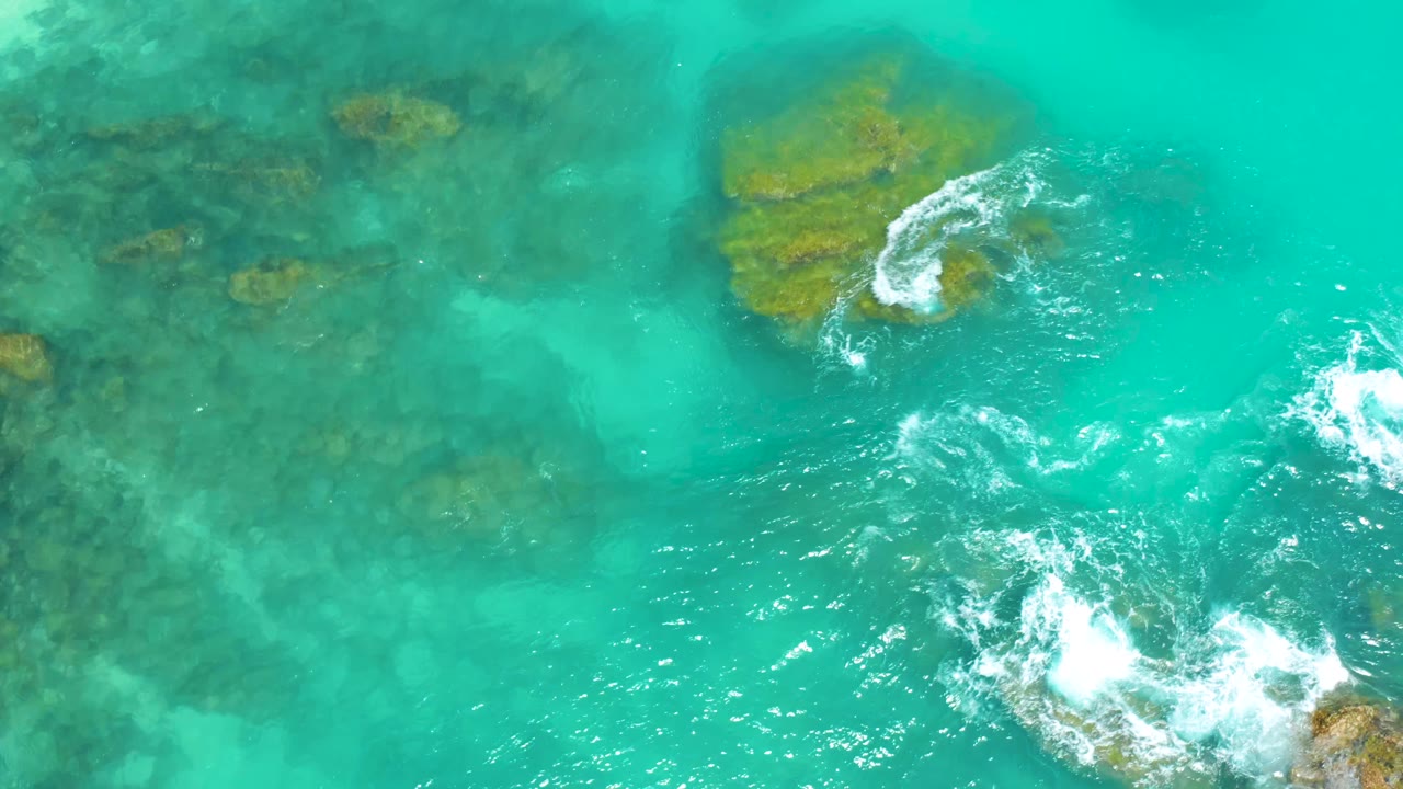 Aerial view at Dreams Beach in Baja California Sur, where blue-green water reveals reef rocks below and white foam gathers along the lower-right rocks, with no visible people.