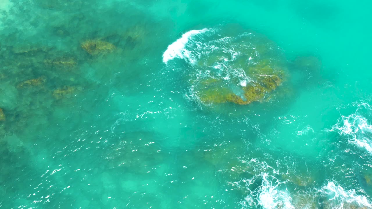 Bright turquoise water at Dreams Beach in Baja California Sur, with a shallow reef rock near center and white foam breaking across its upper edge, with no visible people.