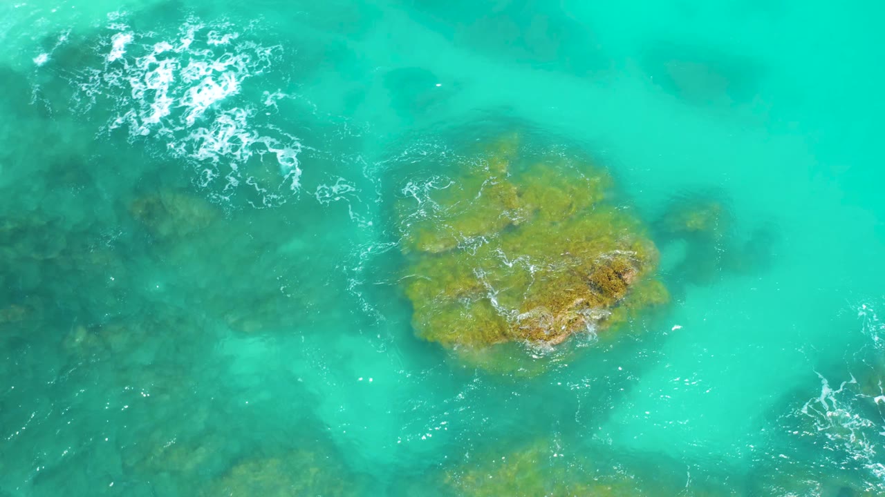 Aerial frame at Dreams Beach in Baja California Sur with the ochre rock slightly right of center, dark rock patches below the surface, pale foam at upper left, and no visible people.