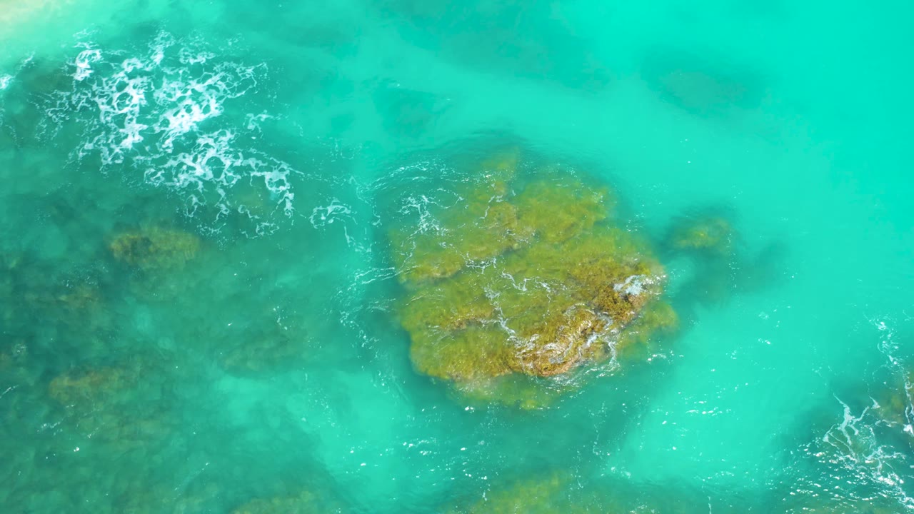 Top-down Dreams Beach frame in Baja California Sur where bright aqua water surrounds a large ochre rock, with darker underwater stone to the left, foam at the upper left and right edge, and no visible people.
