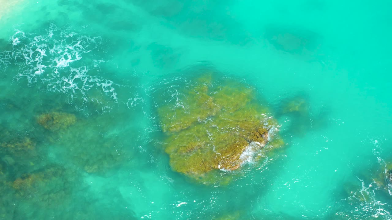 Aerial view at Dreams Beach in Baja California Sur with the main ochre rock lower center, darker rock shelves visible on the left, thin white foam on the rock edge, and no visible people.