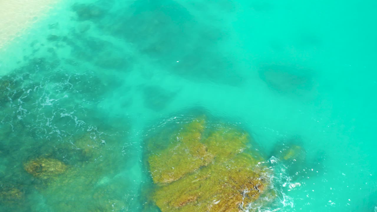 Overhead view at Dreams Beach in Baja California Sur, Mexico, showing turquoise water over dark submerged patches and golden rock near the lower edge, with no visible people.