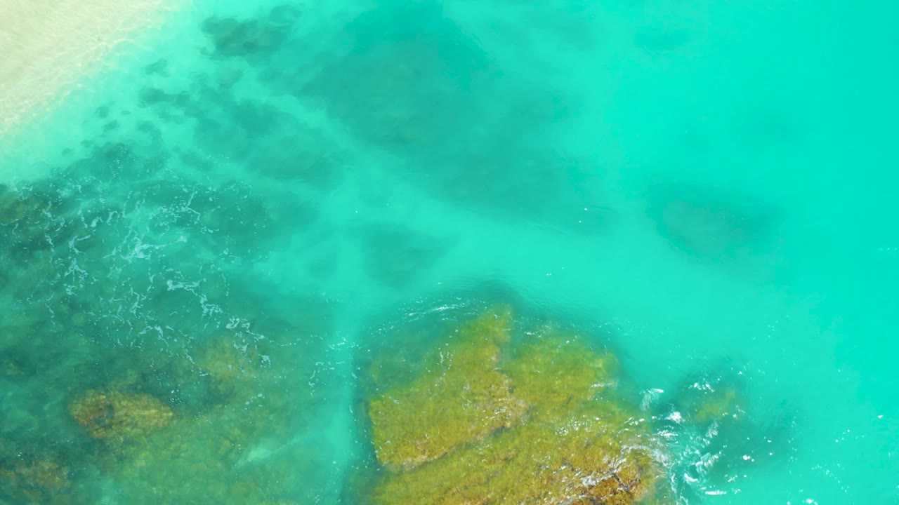 Aerial view at Dreams Beach in Baja California Sur, Mexico, showing bright turquoise sea around a golden rock patch, darker underwater areas to the left, and no visible people.