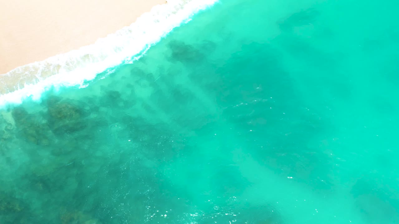 Aerial frame from Dreams Beach in Baja California Sur, Mexico, showing a brighter white foam band along the upper shoreline and clear turquoise water with darker underwater patches, with no visible people.
