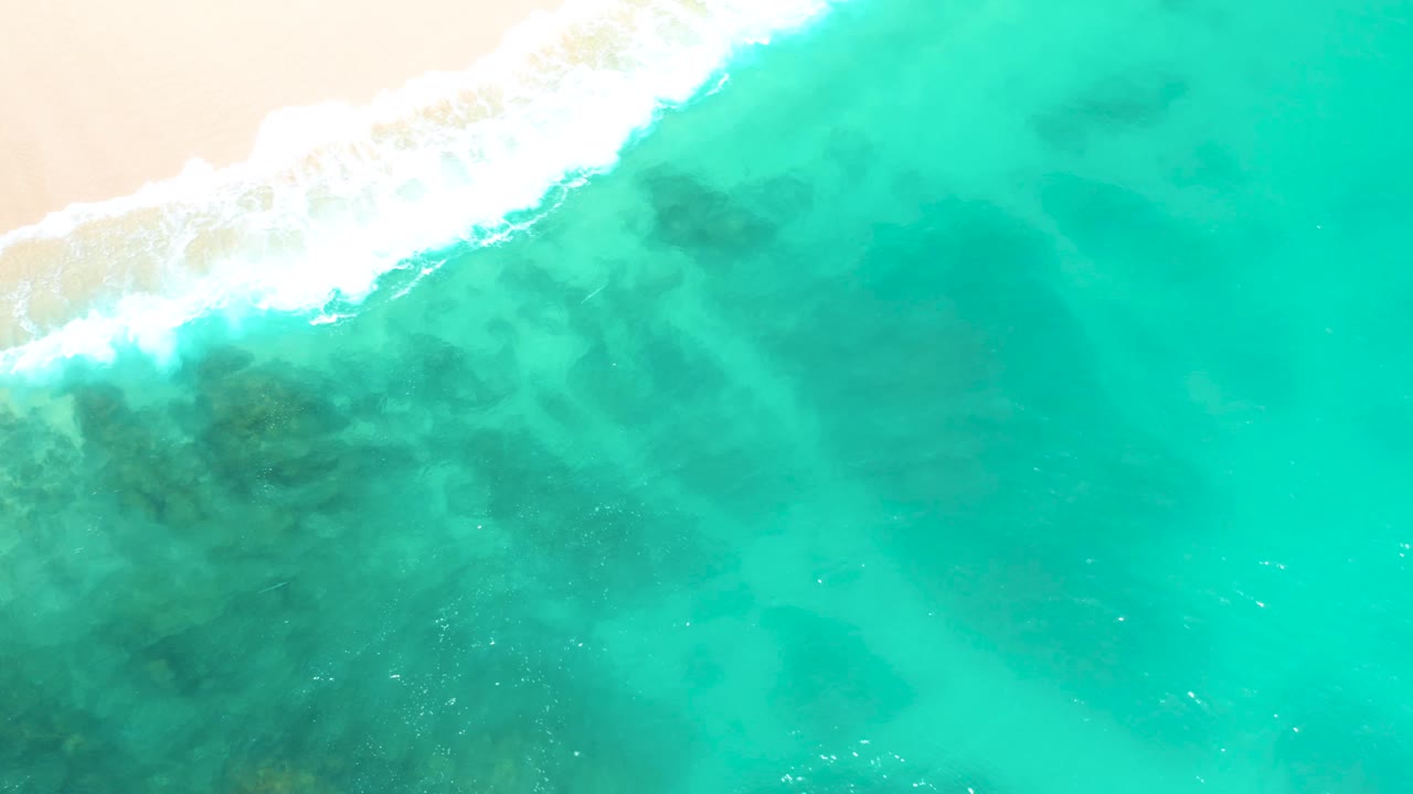 Steep overhead view at Dreams Beach in Baja California Sur, Mexico, with surf spread across the top edge, sandy beach just beyond it, and broad turquoise water below, with no visible people.