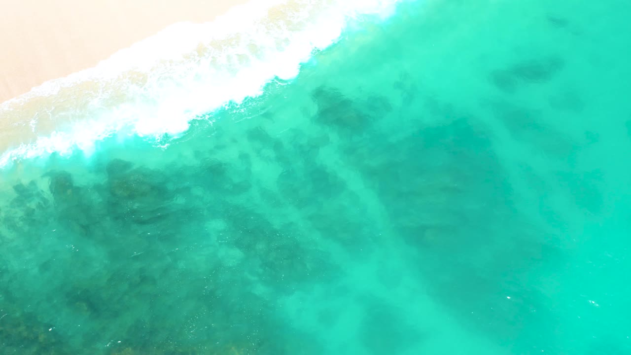 Aerial view at Dreams Beach in Baja California Sur, Mexico, where a foamy wave reaches diagonally from the upper left across pale sand into green-blue water, with no visible people.