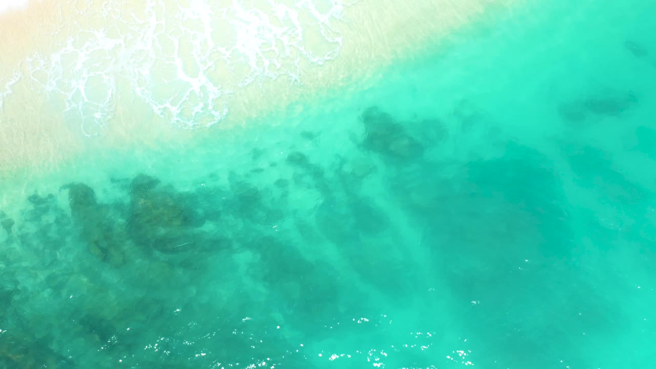 At 0:01, the aerial view keeps pale sand and foamy wash along the upper edge while clear green water reveals a broad dark patch beneath the surface at Dreams Beach, with no visible people.