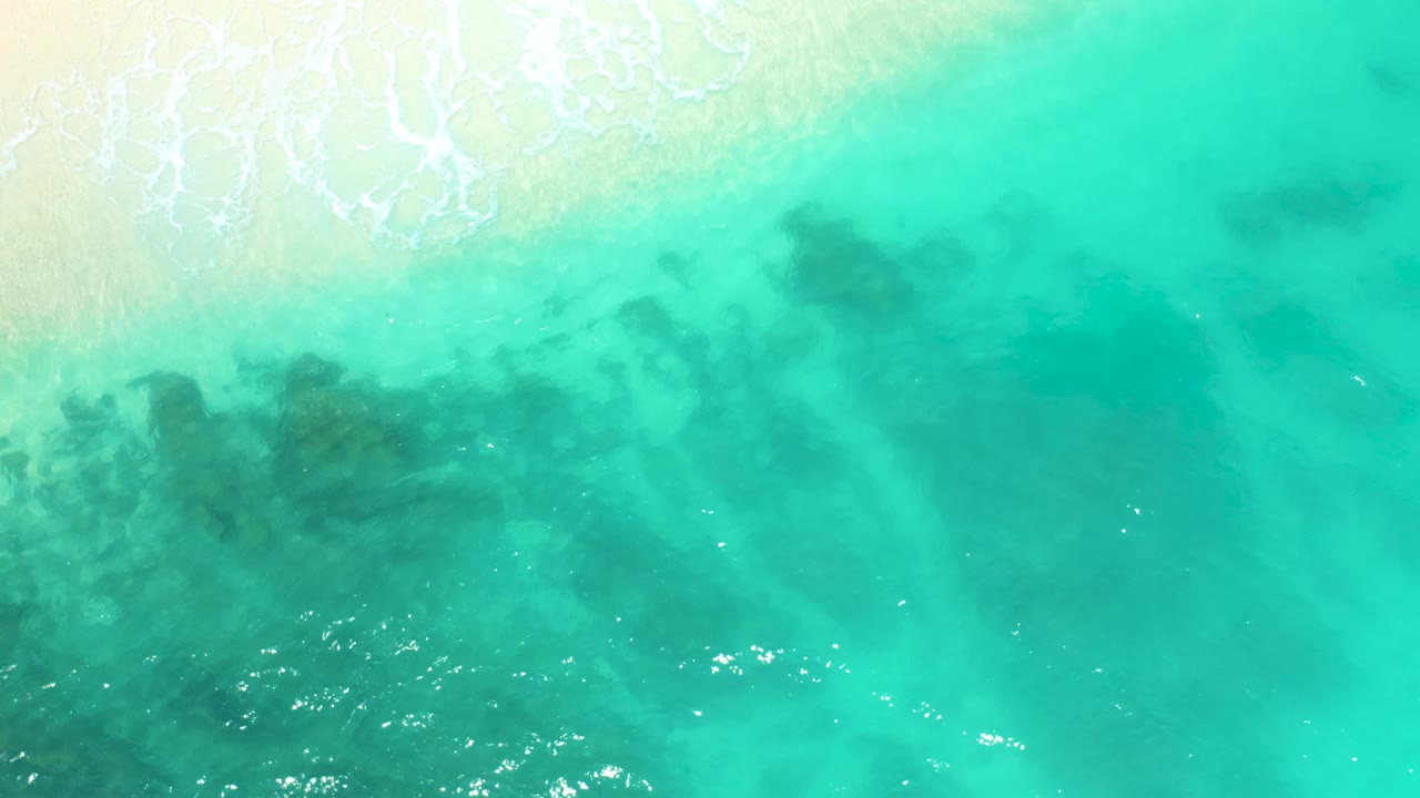 At 0:02, the shore wash sits high in the frame, the turquoise water looks brighter on the right, and darker underwater algae or kelp patches remain visible through the center, with no visible people.