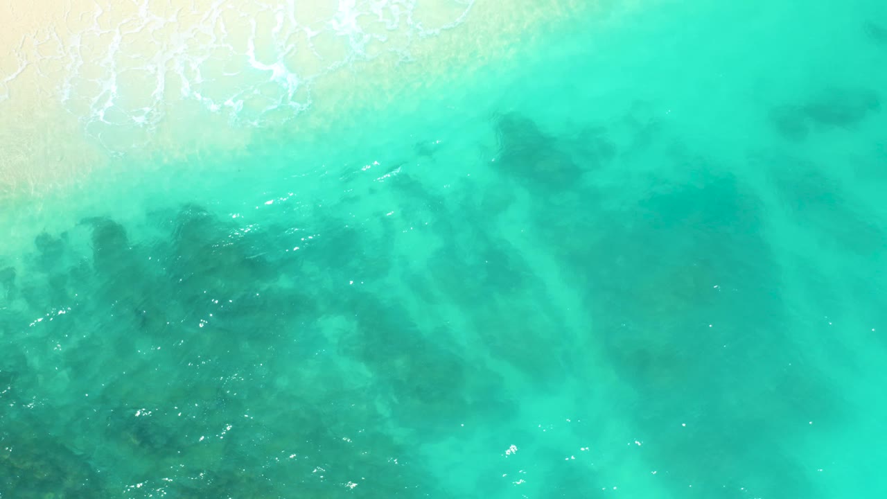 At 0:03, the frame shows pale beach and white wash in the upper left, with clear green water spreading across the view and darker submerged material clustered lower left, no visible people.