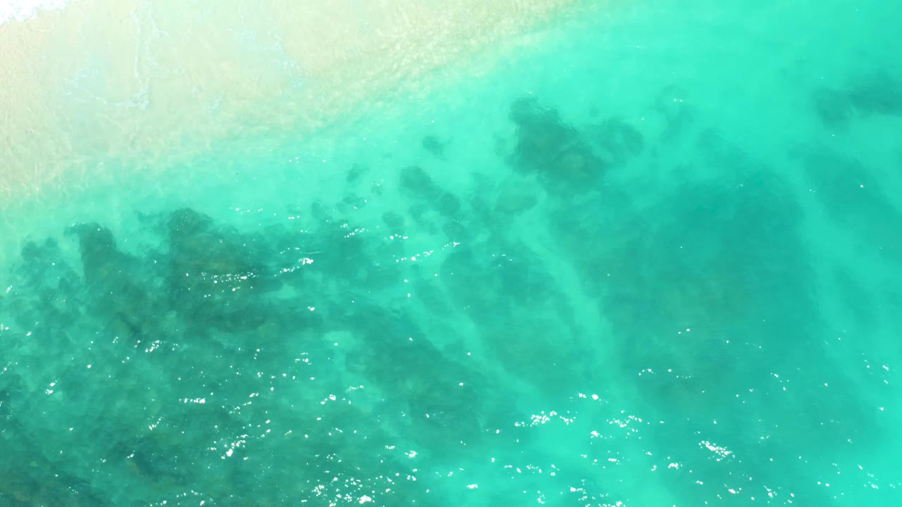 Overhead frame of bright aqua sea at Dreams Beach in Baja California Sur, Mexico, with dark underwater shapes, scattered light reflections, and no visible people.