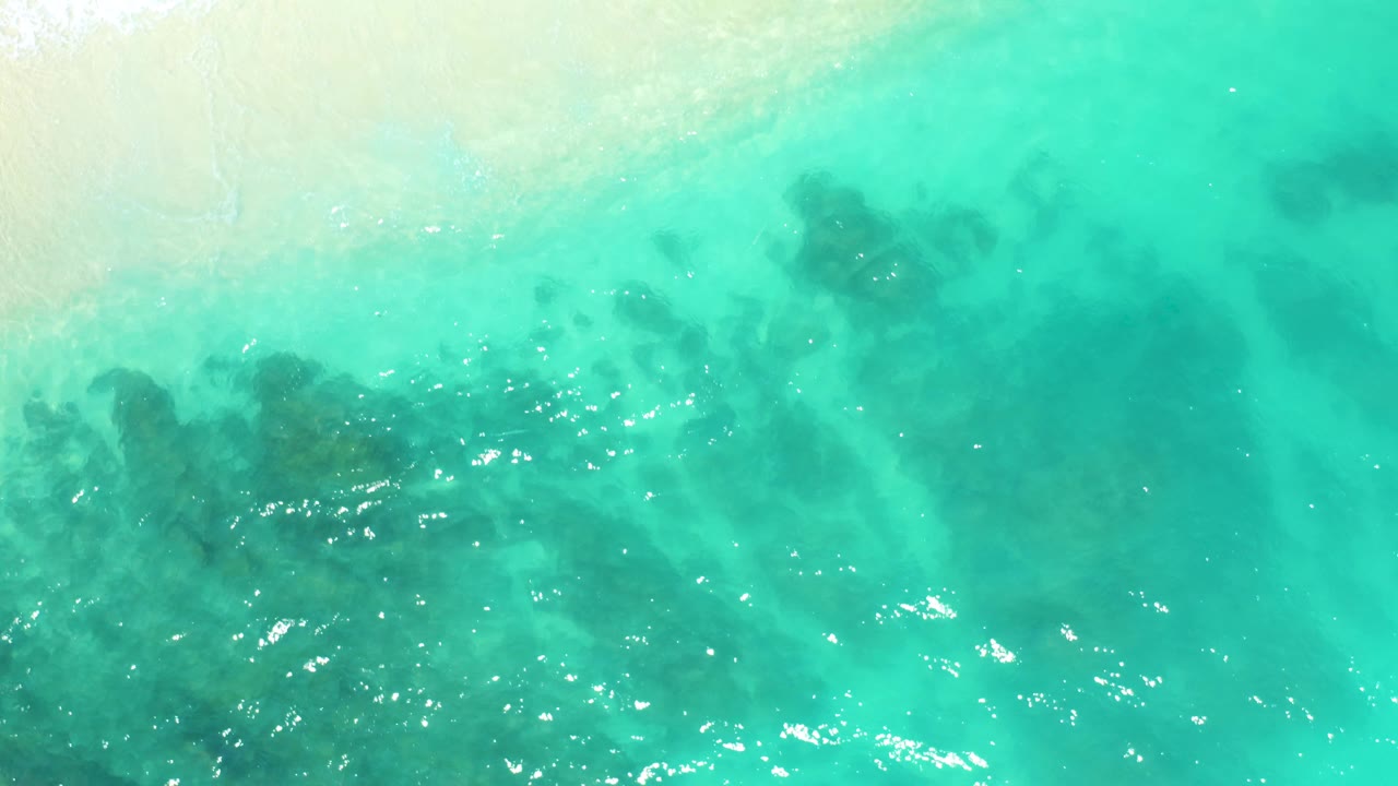 Top-down turquoise sea at Dreams Beach in Baja California Sur, Mexico, with darker submerged areas spread across the frame, bright ripples, and no visible people.