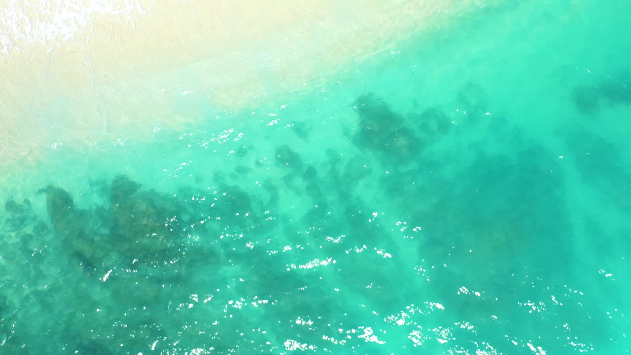 Overhead view at Dreams Beach in Baja California Sur, Mexico, where pale sand enters the upper frame above turquoise shallows and dark submerged patches, with no visible people.
