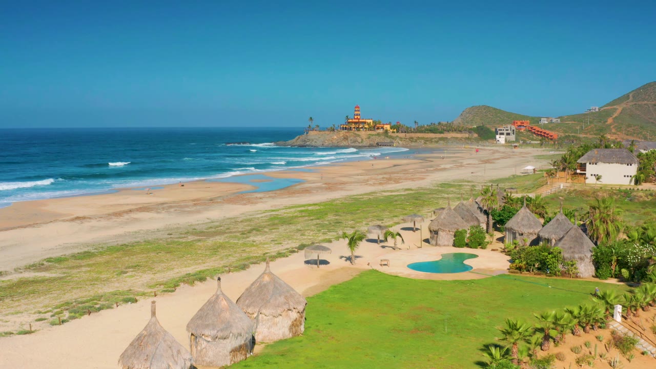 Wide elevated view in Cerritos, Baja California Sur with surf rolling toward a long beach, huts and pool in the lower frame, and no visible people.