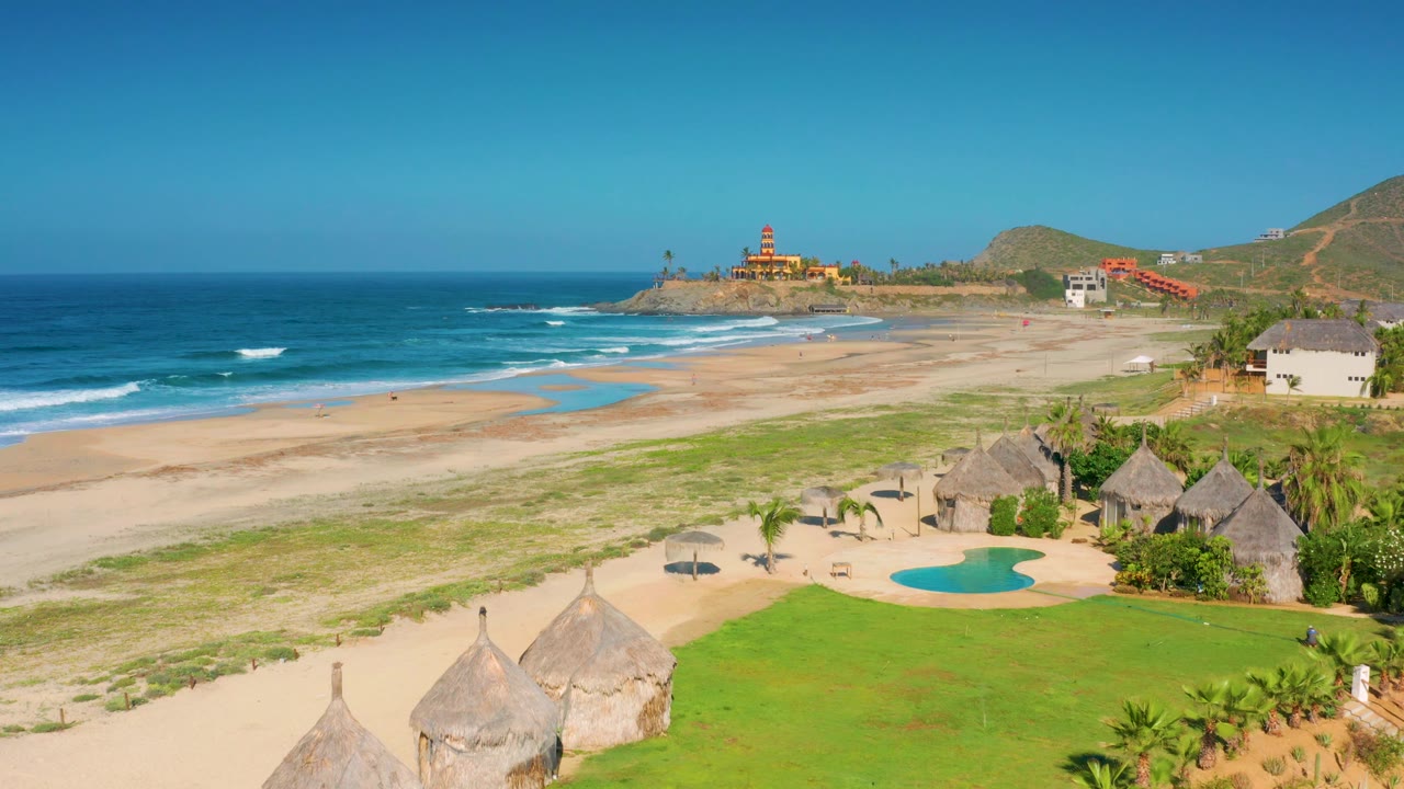 Sunny aerial frame in Cerritos, Baja California Sur with the beach stretching across the center, a small pool and huts at lower right, and no visible people.