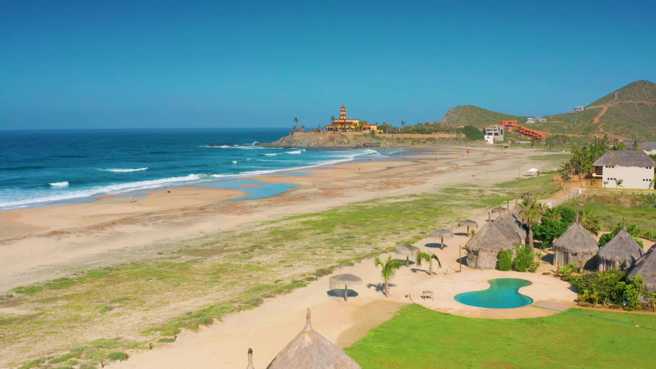 Aerial frame over Cerritos with the wave line slightly shifted, the sandy beach stretching across the middle, huts and pool at lower right, distant hills, and no visible people.