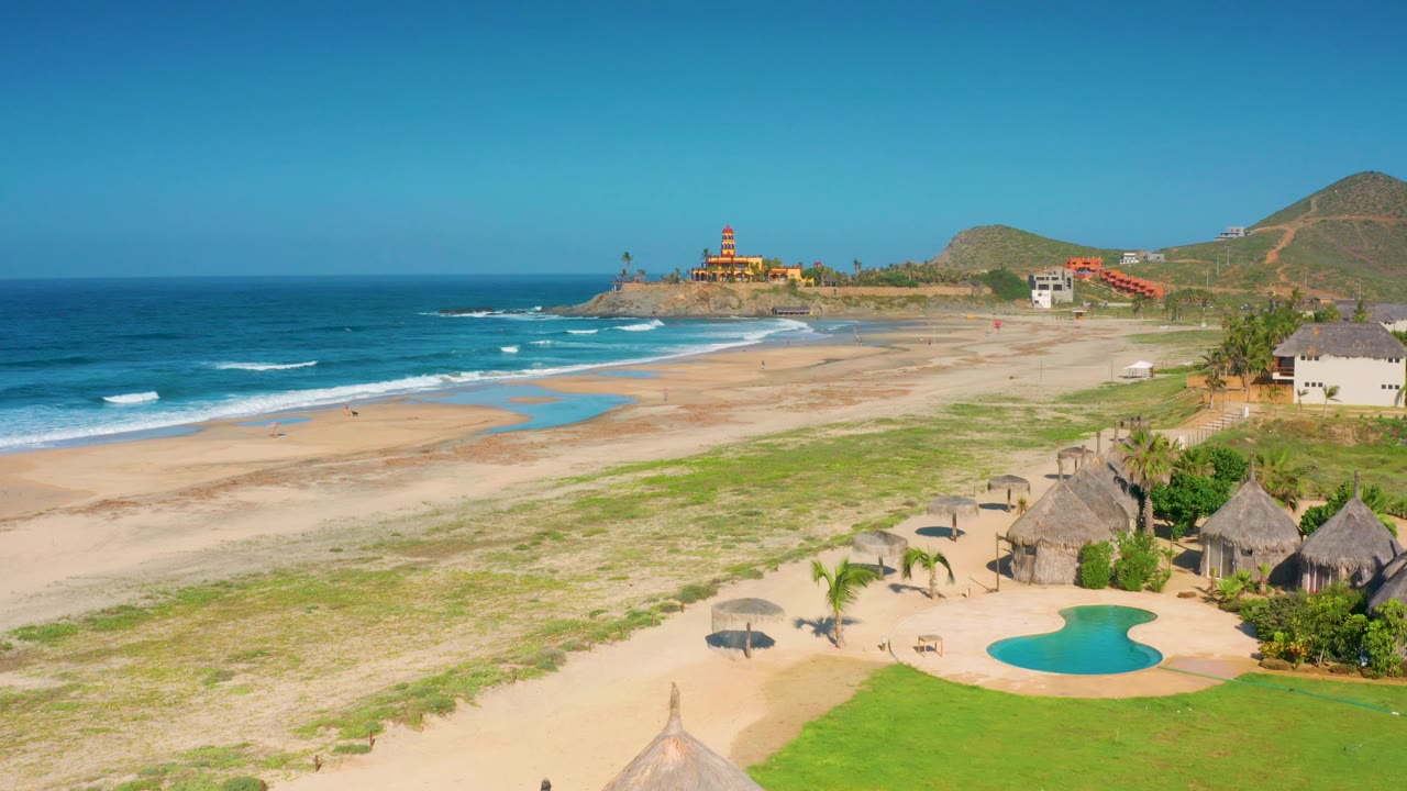 Third aerial frame from Cerritos showing a bright blue sea, small waves, open sand, the pool and thatched roofs at the lower right edge, a hillside beyond the beach, and no visible people.